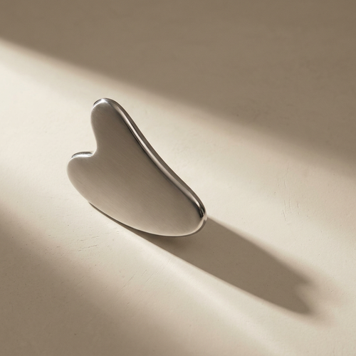 Lumi Cryo Gua Sha, brushed stainless steel, on a bone plaster counter in window light