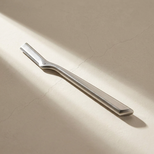 Lumi Editorial Dermaplaning Tool, stainless steel, on a bone plaster counter in window light