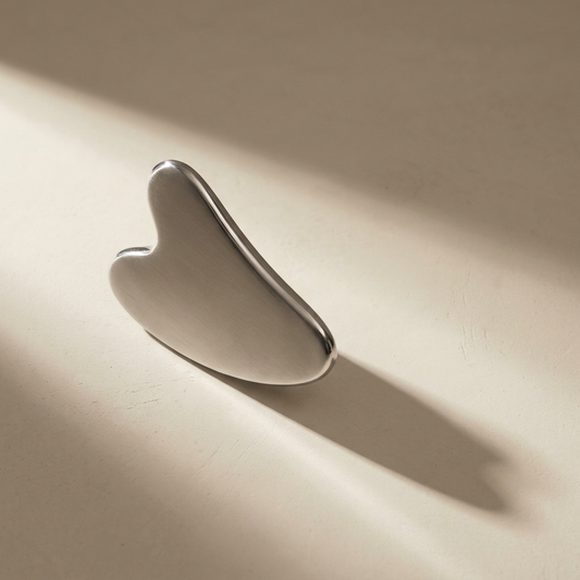 Lumi Cryo Gua Sha, weighted stainless steel, on a bone plaster counter in window light